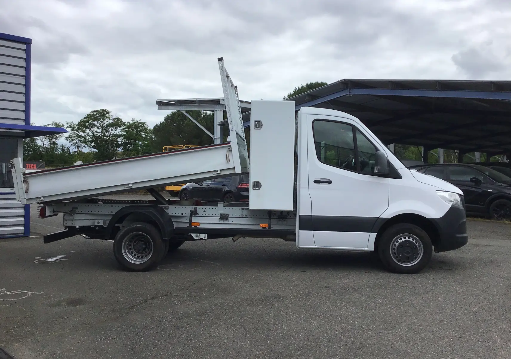 Vue de profil côté droit d'un Mercedes Sprinter blanc avec benne relevée et coffre latéral blanc.