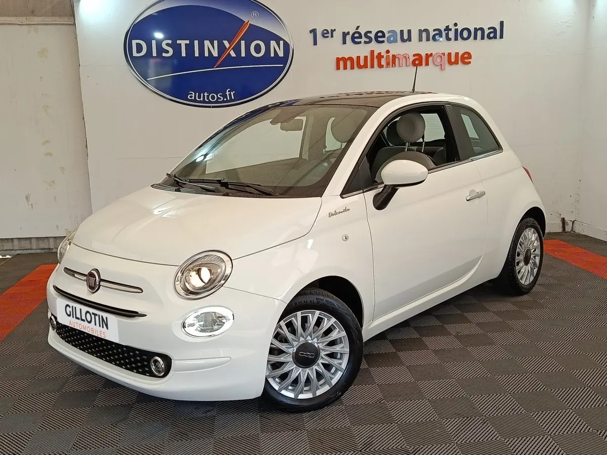 Vue 3/4 avant droite d'une Fiat 500 Dolcevita blanche, avec jantes multibranches et phares allumés en intérieur showroom.