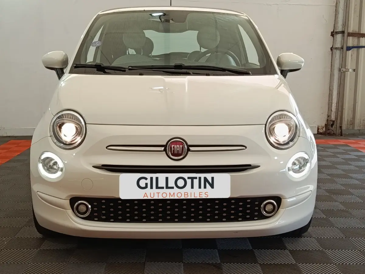 Vue frontale d'une FIAT 500 blanche avec phares allumés et calandre noire à motifs ovales.