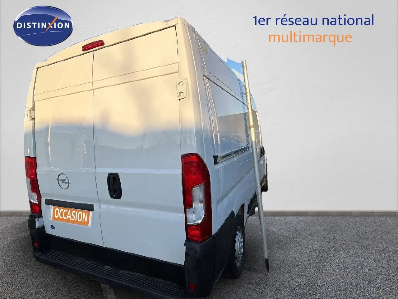 Vue 3/4 arrière droite d’un Opel Movano Fourgon blanc Icy 2023 avec porte arrière fermée et panneau publicitaire à côté.
