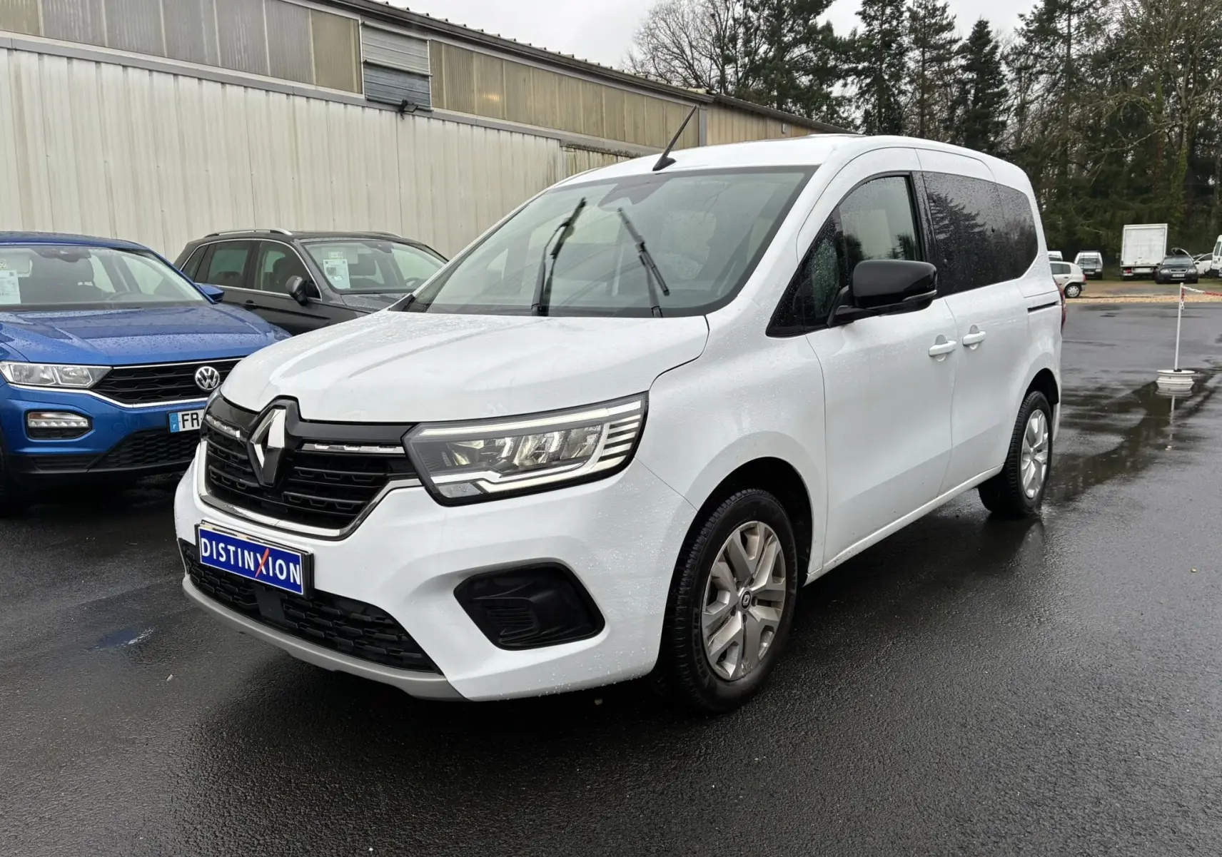 Renault Kangoo TCE 100 Zen blanc vu en 3/4 avant droit sur parking humide, avec calandre noire et phares LED.