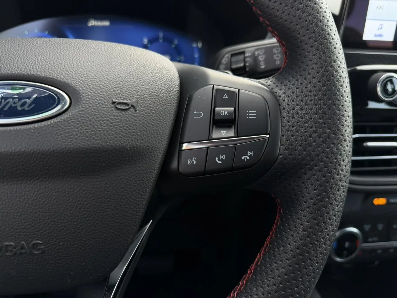 Gros plan sur le volant perforé noir avec surpiqûres rouges du Ford Kuga 2023, boutons de commande multifonctions visibles.