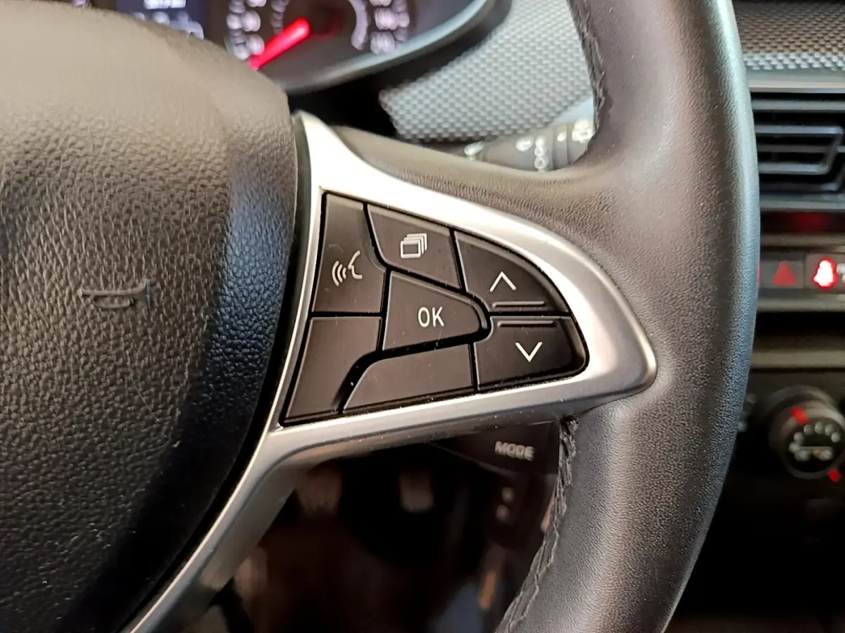 Gros plan sur les commandes multifonctions noires et argentées du volant d'une Dacia Sandero blanche, tableau de bord en arrière-plan.