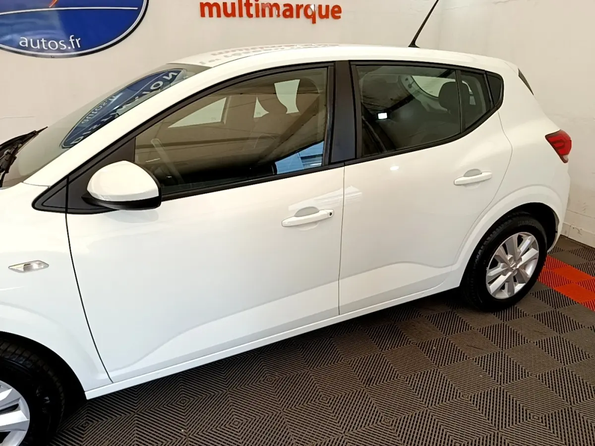 Vue latérale droite d'une Dacia Sandero Confort TCe 90 blanche, en intérieur sur sol à motifs géométriques.