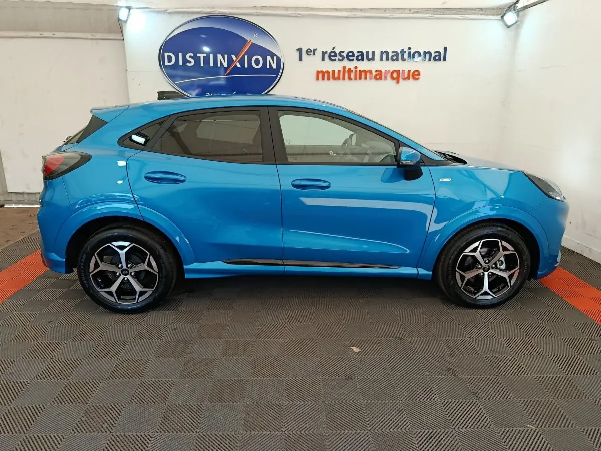 Vue latérale gauche d'un Ford Puma bleu 2025 ST-LINE avec jantes alliage et vitres teintées en intérieur showroom.