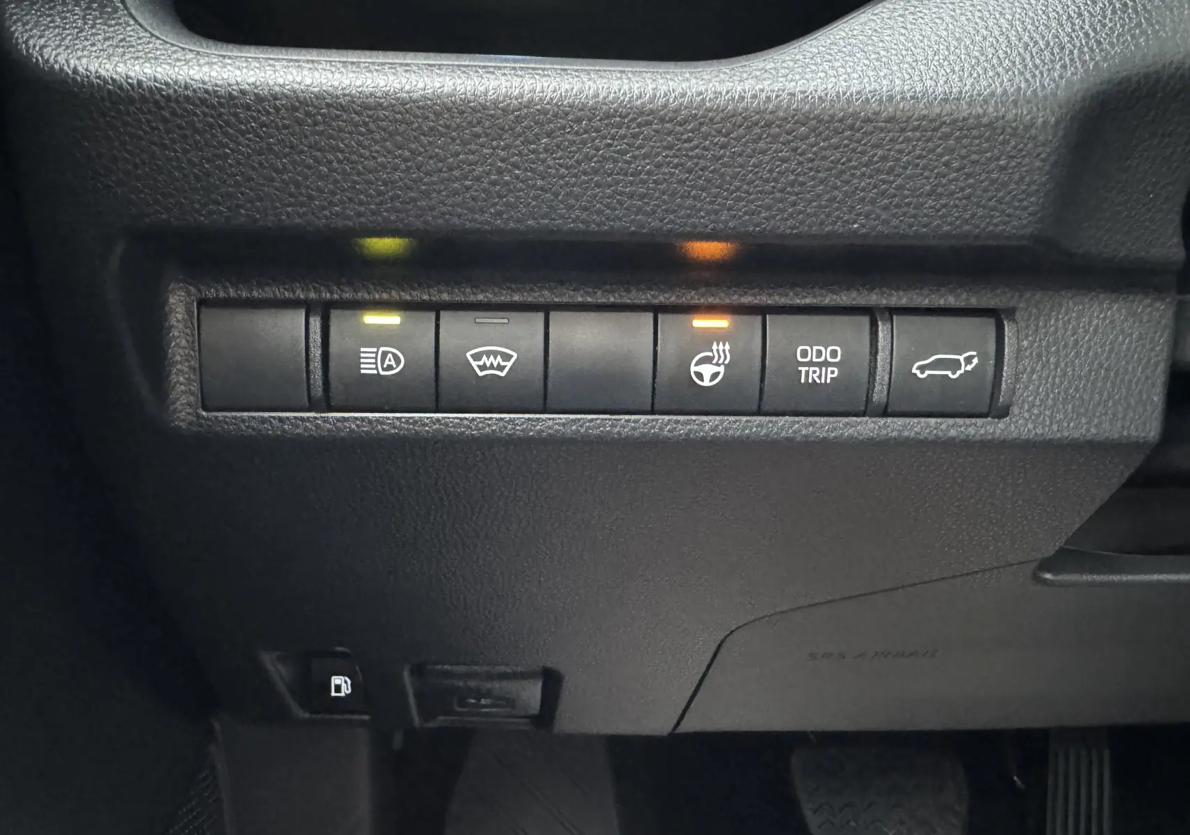 Gros plan sur les commandes du tableau de bord du Suzuki Across blanc 2023, avec voyants allumés pour phares et volant chauffant.