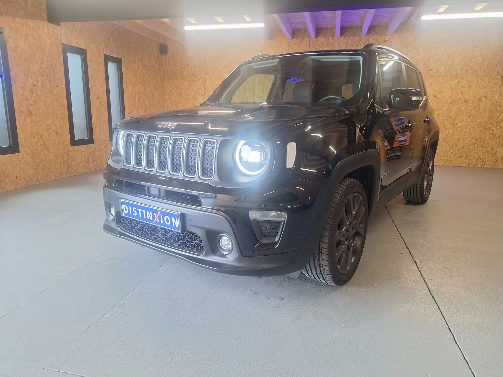 Jeep Renegade noir en 3/4 avant droit, phare allumé, posé dans un intérieur avec murs en bois clair.
