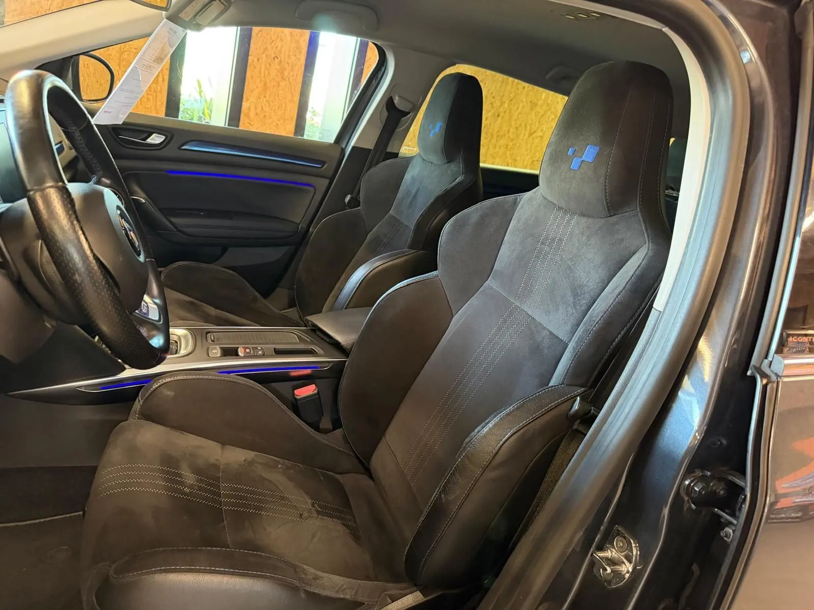 Vue intérieure côté conducteur de la Renault Mégane Break GT 2017, sièges sport noirs avec surpiqûres et logo bleu.