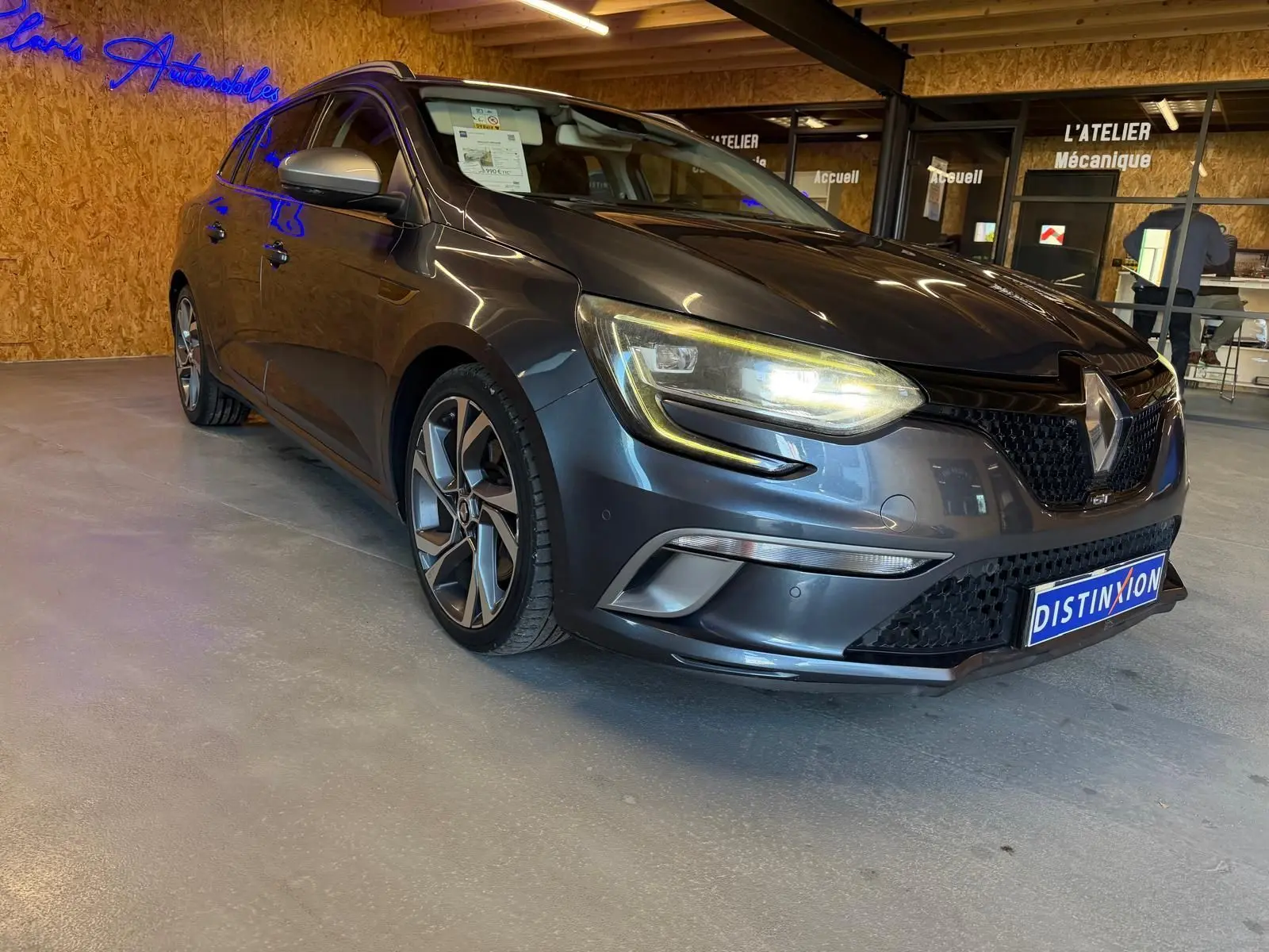 Renault Mégane Break GT gris en 3/4 avant droit, phares allumés, jantes alliage et intérieur visible en showroom.