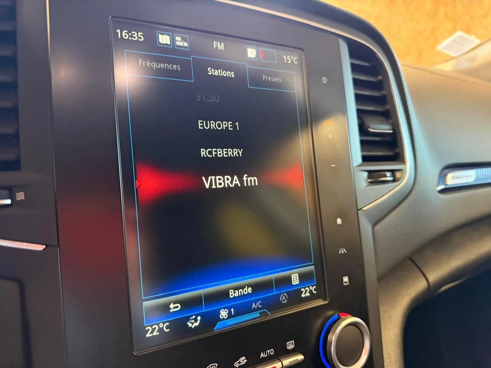 Écran tactile central du tableau de bord de la Renault Mégane Break gris, affichant les stations radio FM avec climatisation à 22°C.