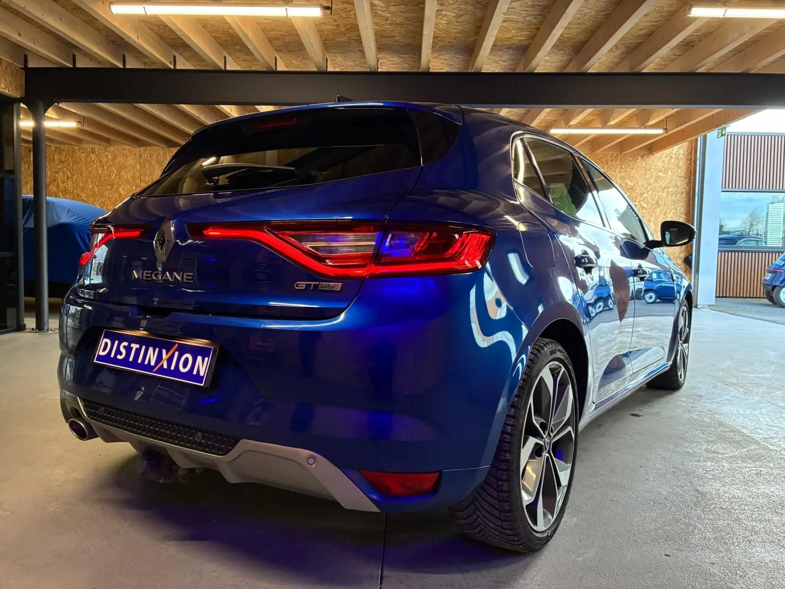 Vue 3/4 arrière droite d'une Renault Mégane GT Line bleu métallisé, avec feux arrière allumés, stationnée en garage.