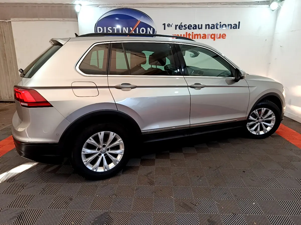 Profil droit d'un Volkswagen Tiguan gris clair 2020 avec jantes alliage et barres de toit noires dans un showroom