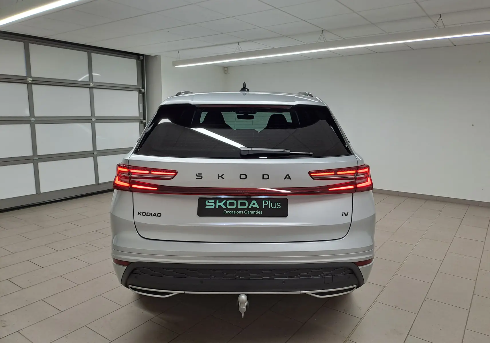 Vue arrière d'un Skoda Kodiaq gris argent 2025 avec feux LED allumés et crochet d'attelage pivotant en intérieur showroom.