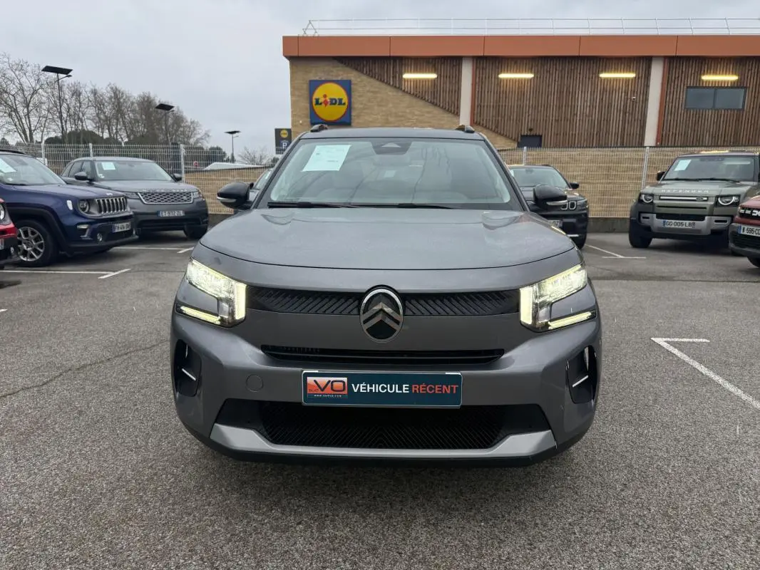 Vue frontale d'une Citroën C3 gris 2025 avec feux allumés et barres de toit visibles sur un parking.