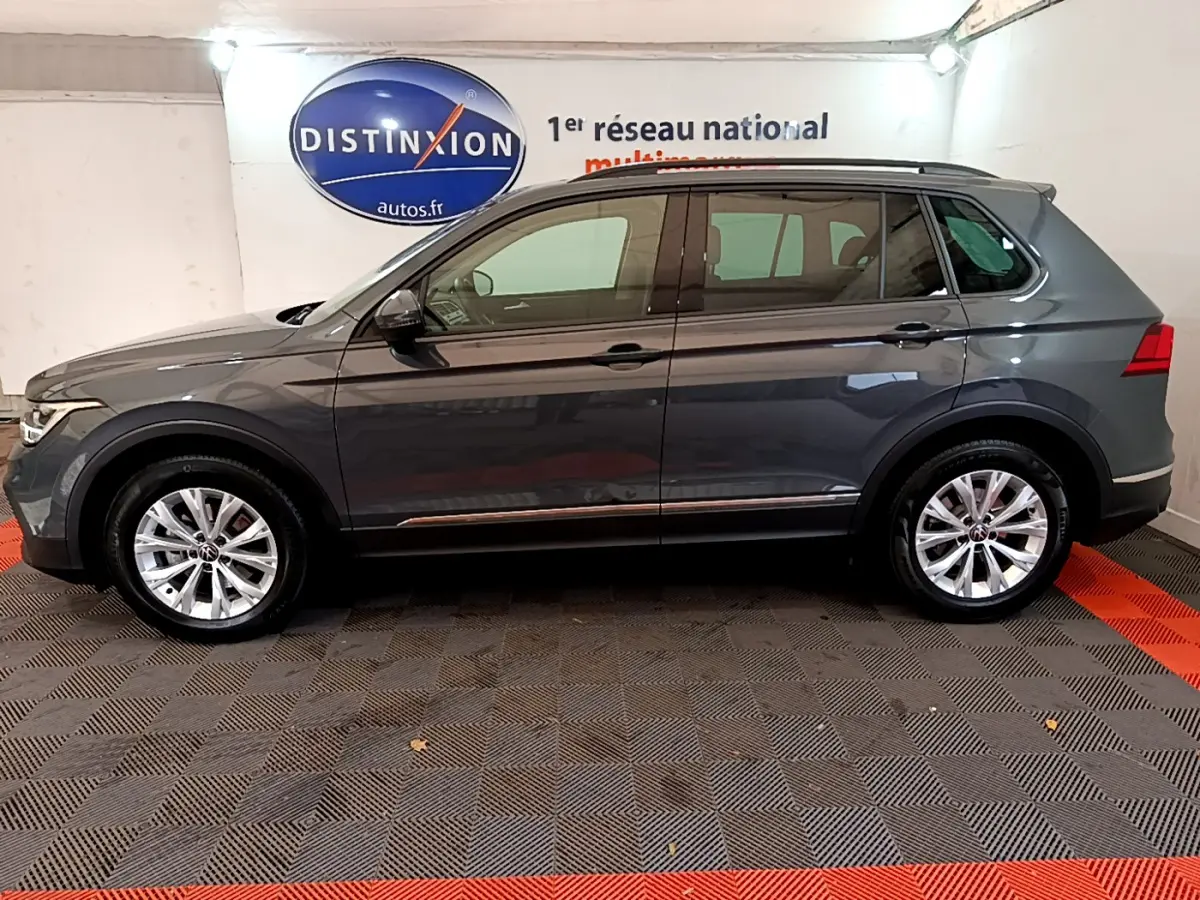 Profil côté gauche d'un Volkswagen Tiguan gris clair 2021 garé en intérieur avec jantes alliage et barres de toit noires.