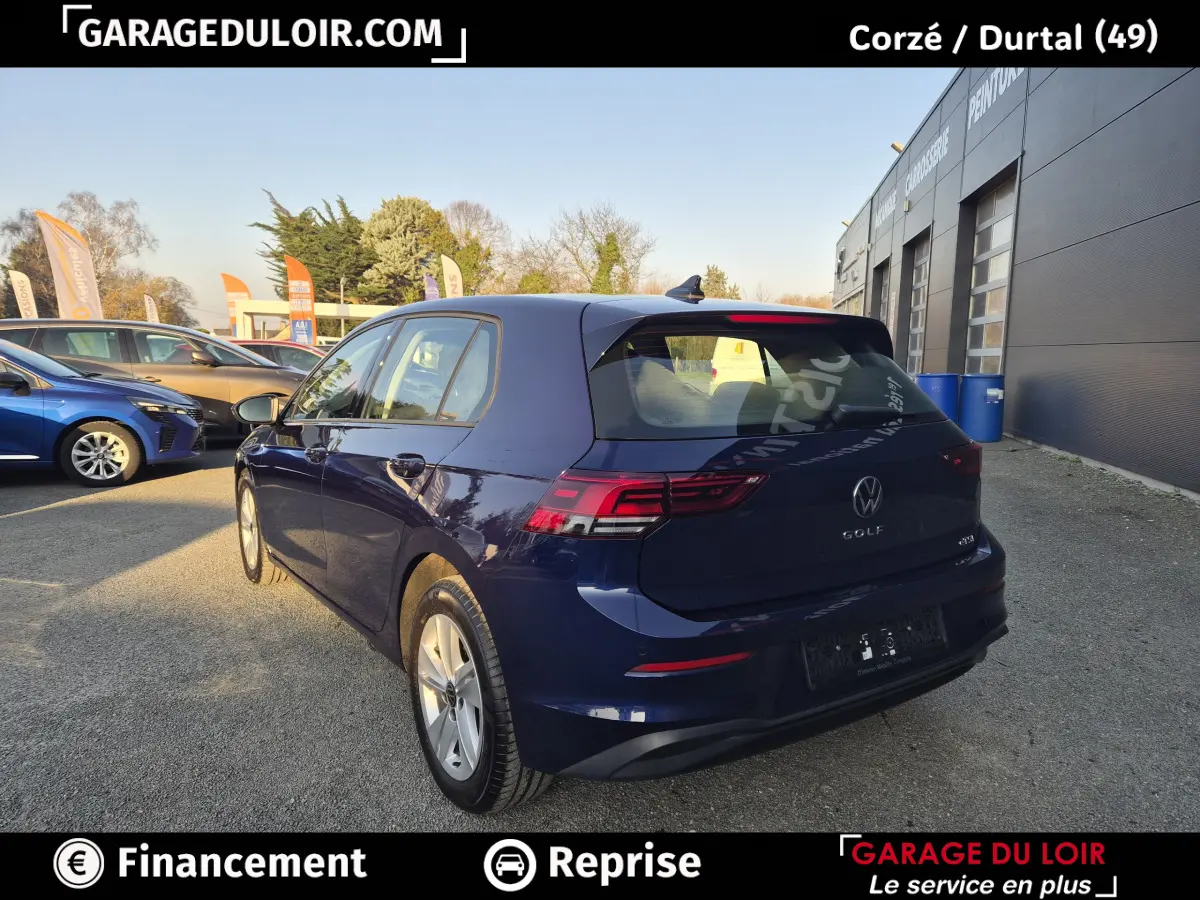 Vue 3/4 arrière droite d'une Volkswagen Golf bleue 2023 stationnée devant un garage, avec feux arrière LED visibles.