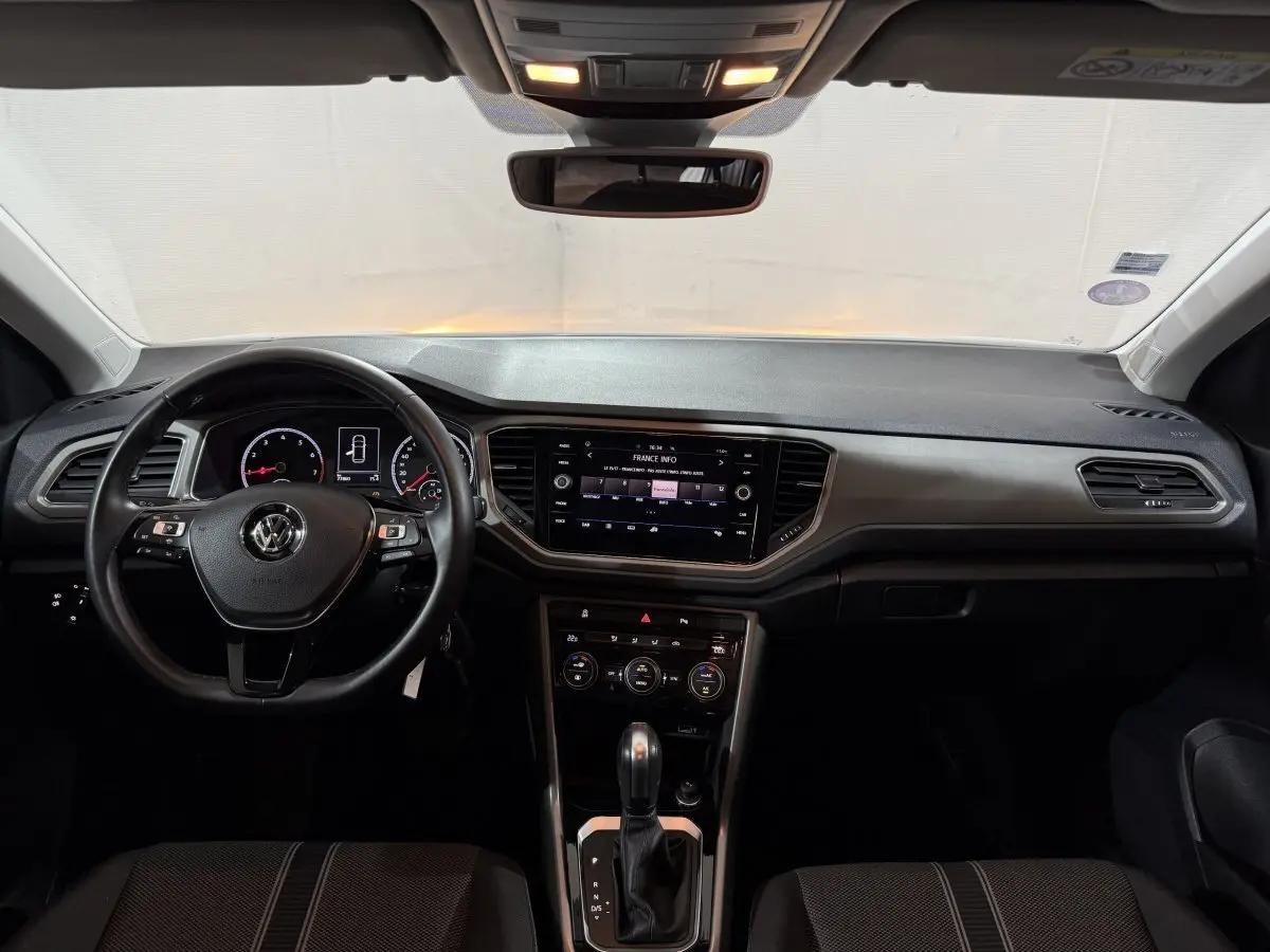 Vue intérieure frontale du tableau de bord et volant cuir noir du Volkswagen T-Roc blanc de 2020 avec écran tactile central allumé.