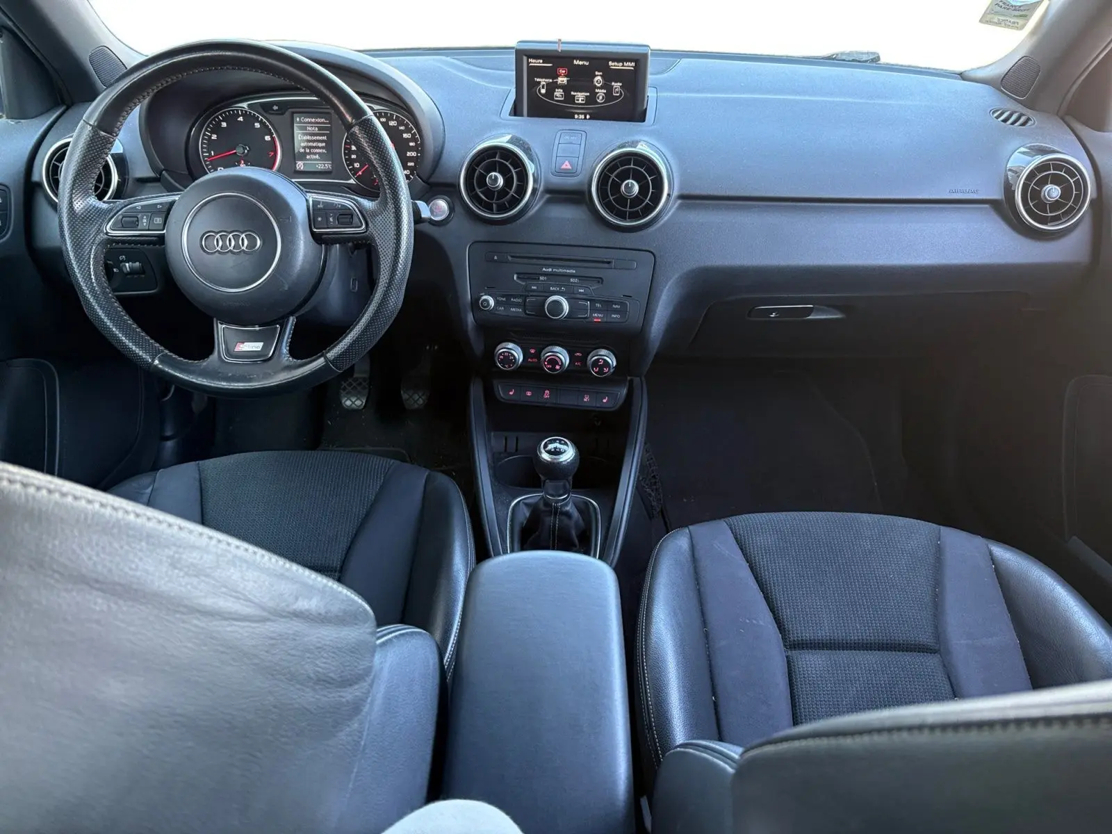 Vue intérieure avant de l'Audi A1 Sportback 1.4 TFSI S-Line 2014, volant sport, écran GPS et levier de vitesses manuel.