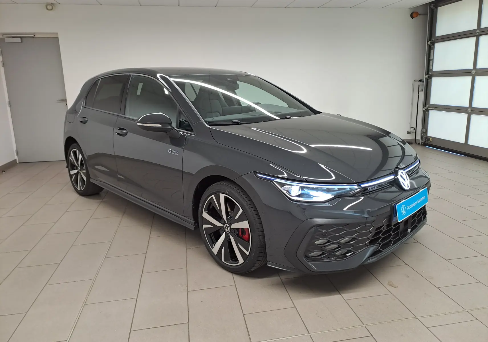 Volkswagen Golf 1.5 eHybrid gris Urano vue 3/4 avant droit, avec badge GTE et jantes sport bicolores.