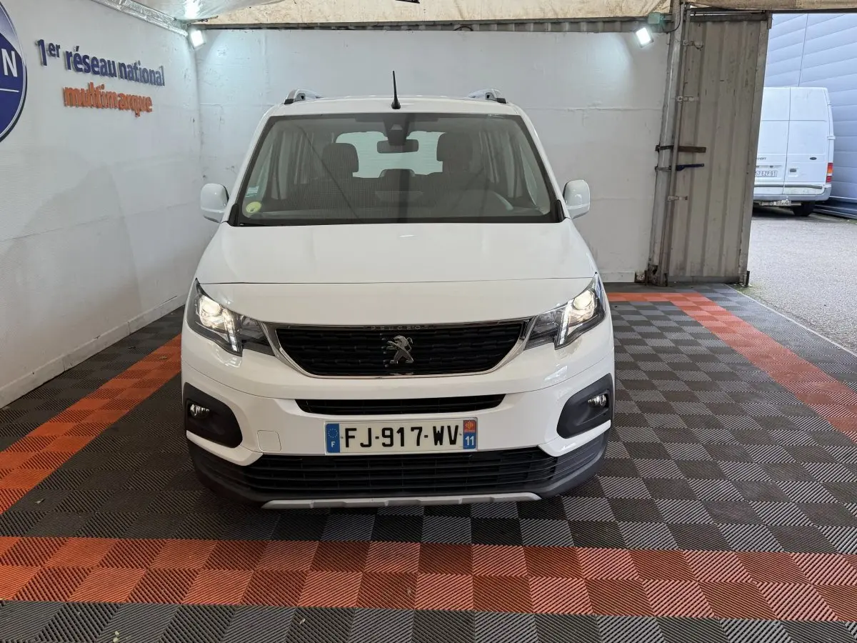 Vue frontale d'un Peugeot Rifter blanc 2019 avec calandre noire et feux allumés dans un garage.