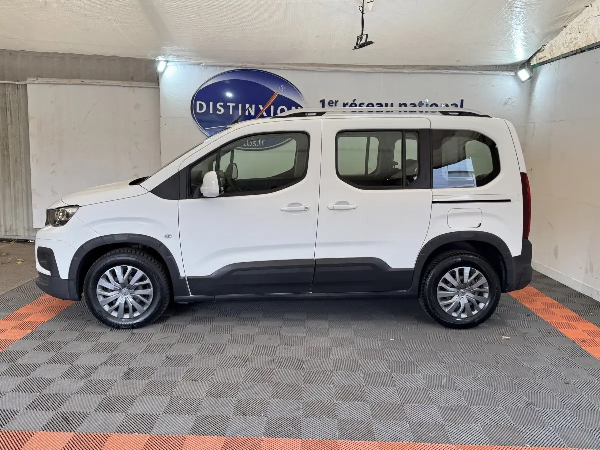 Vue de profil côté gauche d'un Peugeot Rifter blanc 2019 avec protections noires et jantes argentées en intérieur showroom.