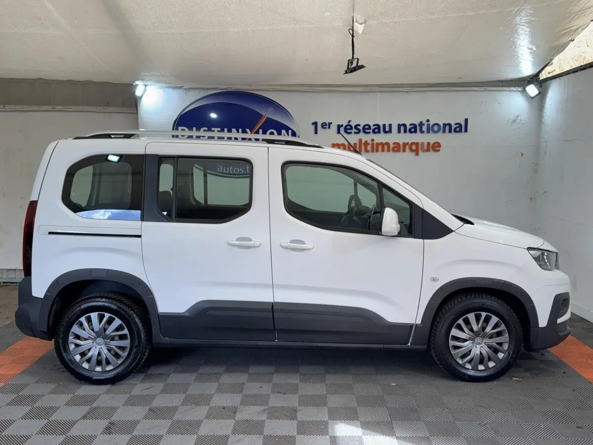 Profil droit d'un Peugeot Rifter blanc 2019 avec jantes alliage et barres de toit chromées en intérieur showroom.