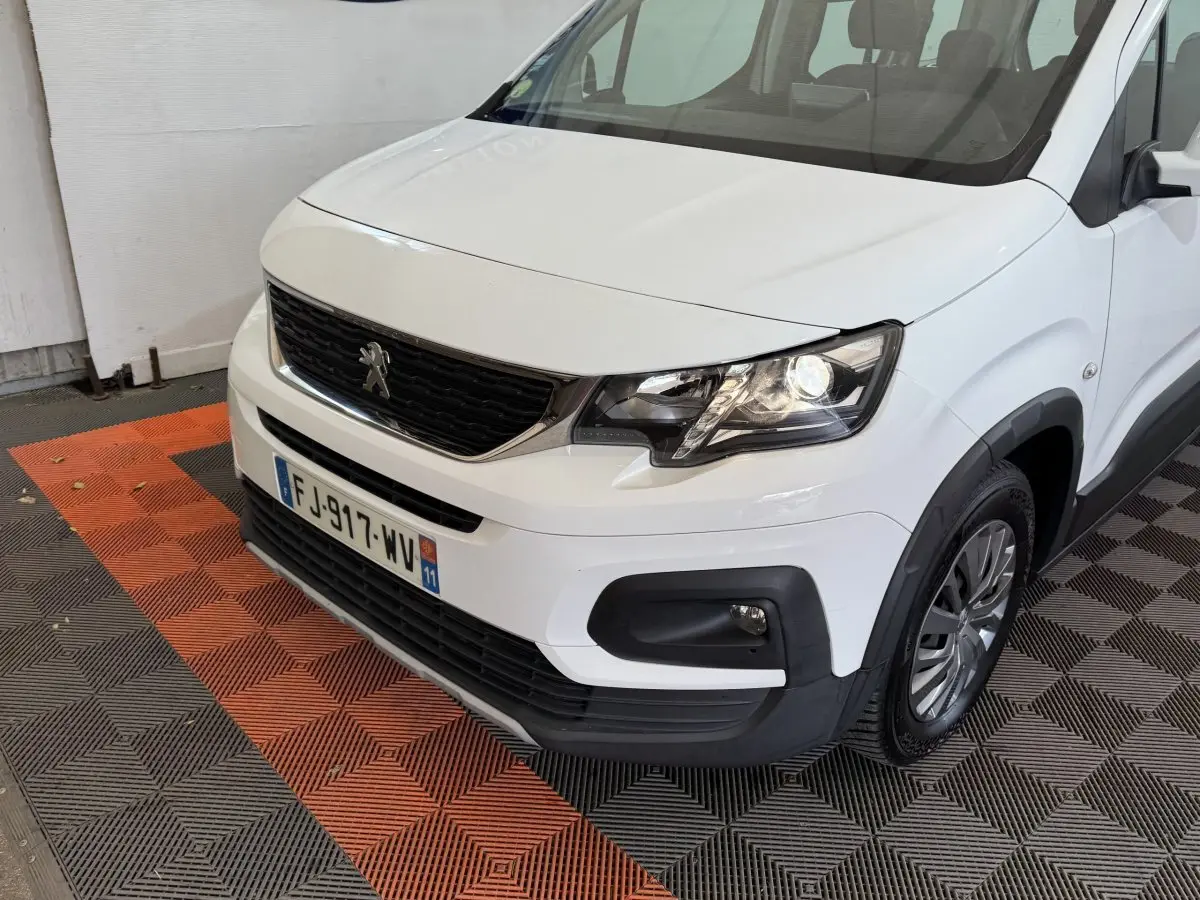 Vue 3/4 avant droit d'un Peugeot Rifter BlueHDi 100 Allure blanc avec calandre noire et feux allumés.