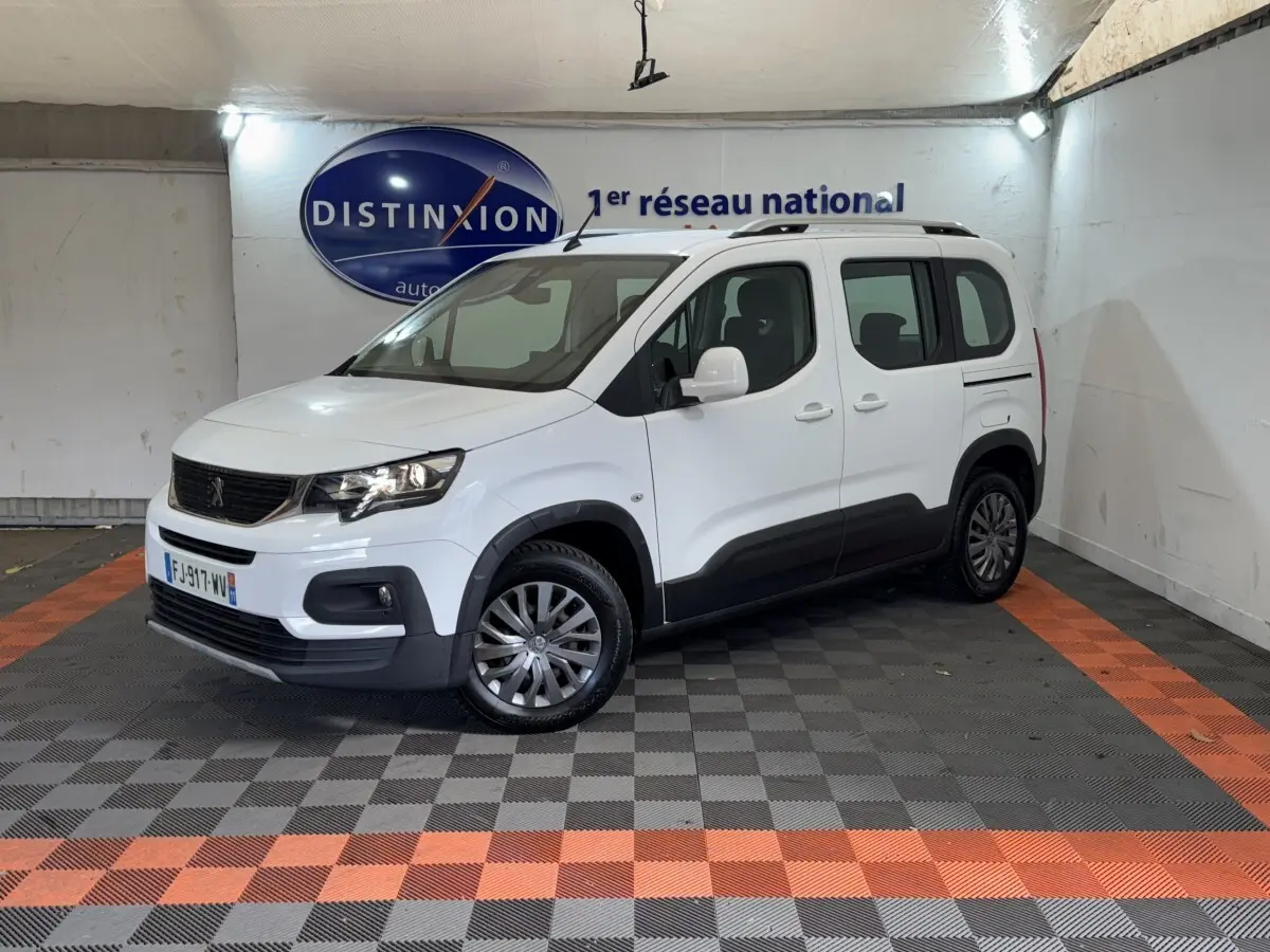 Peugeot Rifter blanc en 3/4 avant droit, avec barres de toit noires et protections latérales noires visibles.