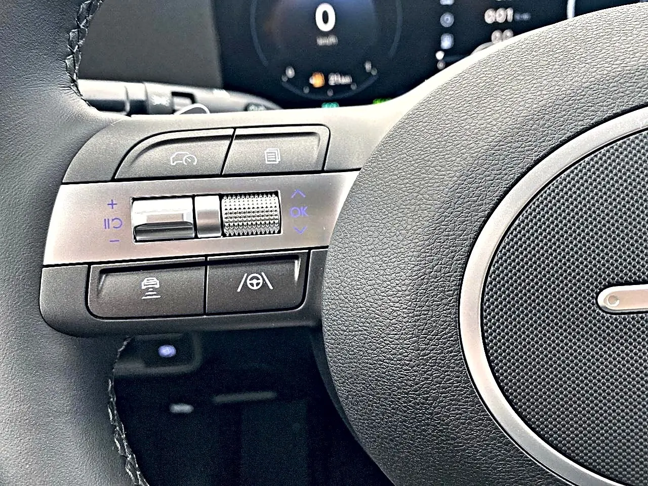 Gros plan sur les commandes à gauche du volant cuir du Hyundai Kona gris, avec tableau de bord numérique en arrière-plan.