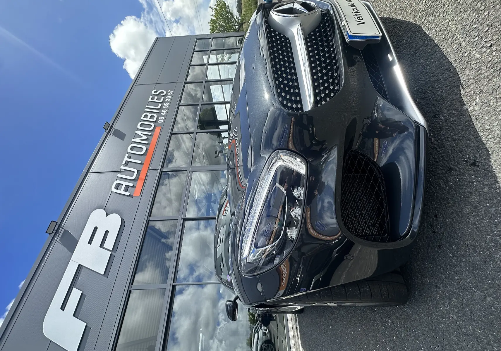 Vue 3/4 avant droit d'une Mercedes AMG GT gris foncé stationnée devant un bâtiment avec logo FB Automobiles.