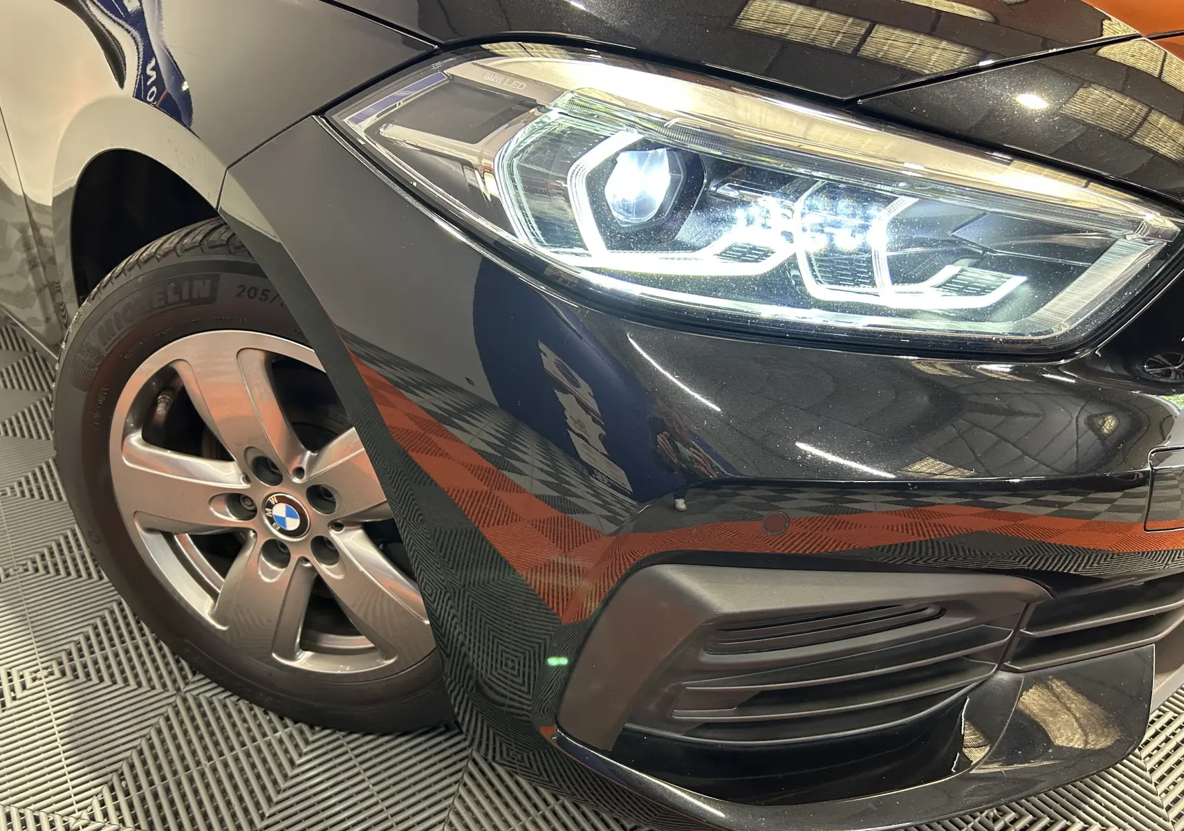 Gros plan sur le phare avant droit allumé et la roue en alliage d'une BMW Série 1 noire 2023.