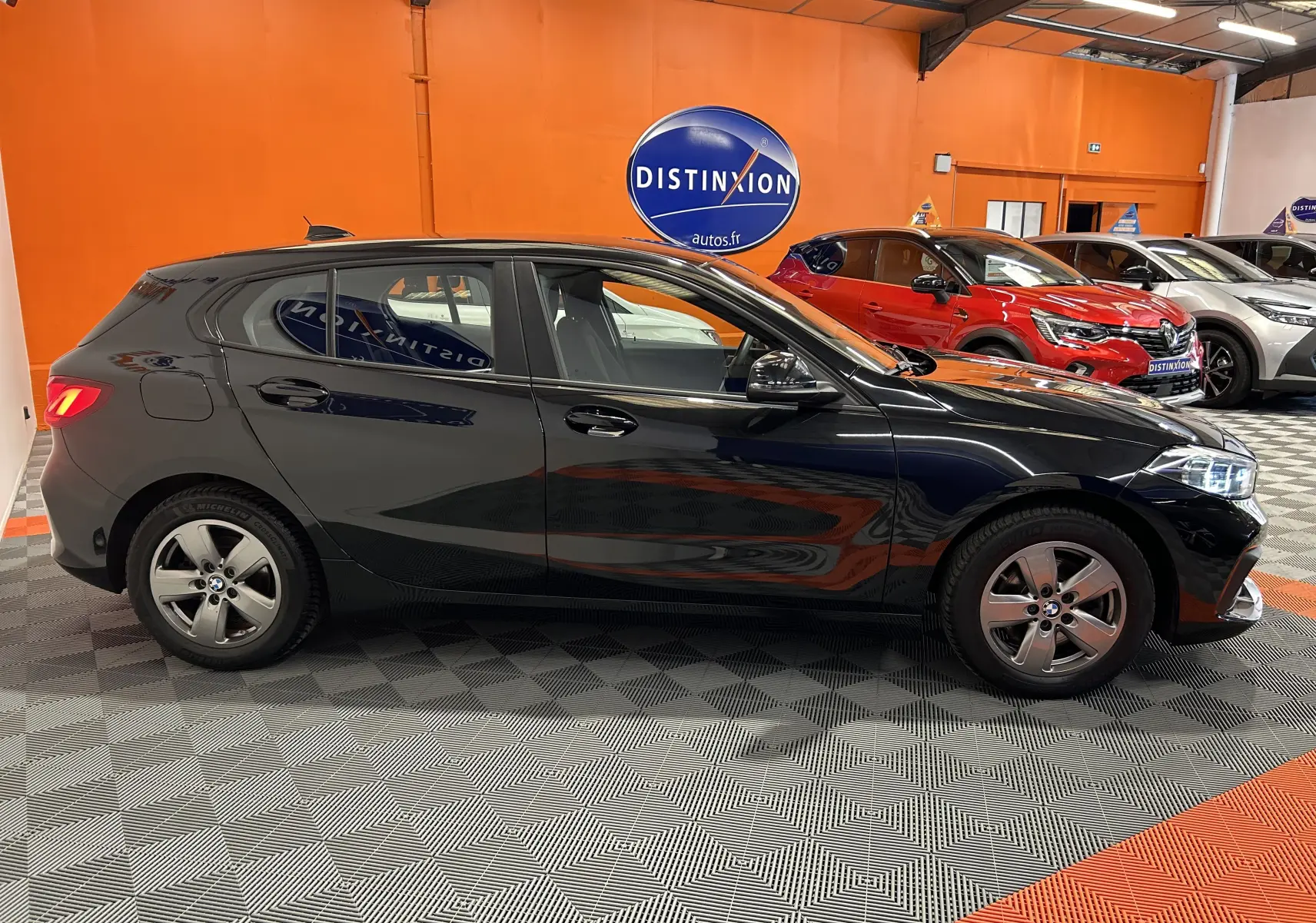 Profil côté gauche d'une BMW Série 1 noire 5 portes de 2023, dans un showroom avec sol à motifs gris et orange.