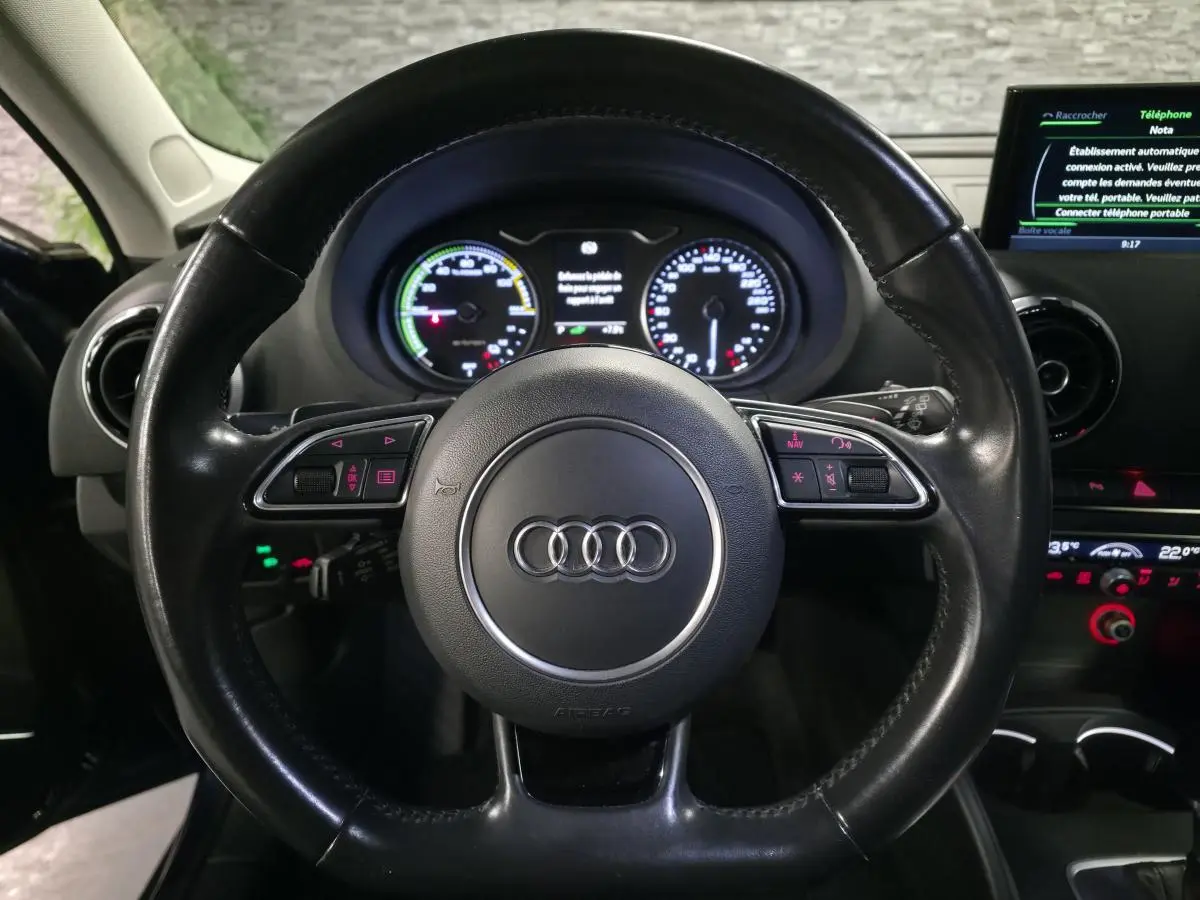 Vue intérieure centrée sur le volant cuir noir de l'Audi A3 Sportback avec tableau de bord et écran multimédia allumé.