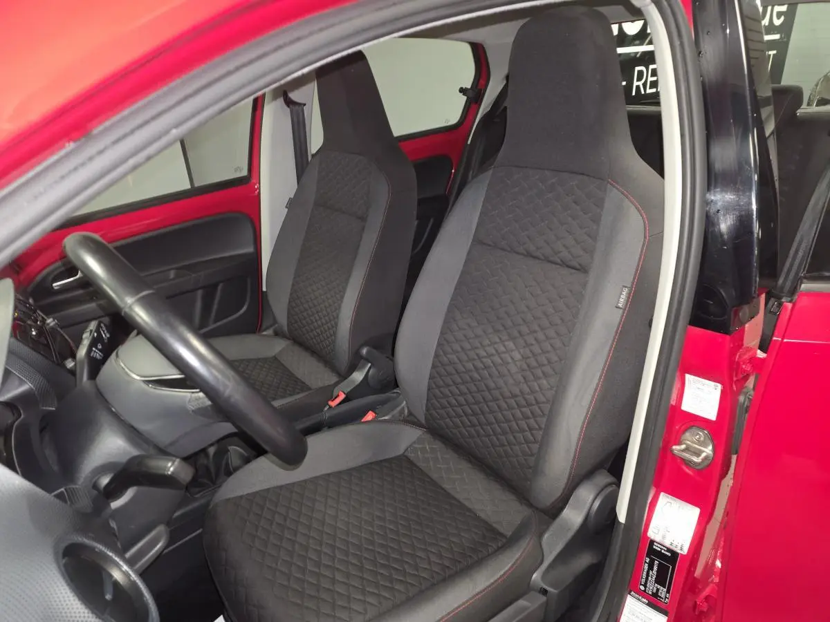 Vue intérieure côté conducteur d'une Volkswagen Up rouge, sièges avant en tissu noir avec surpiqûres rouges.
