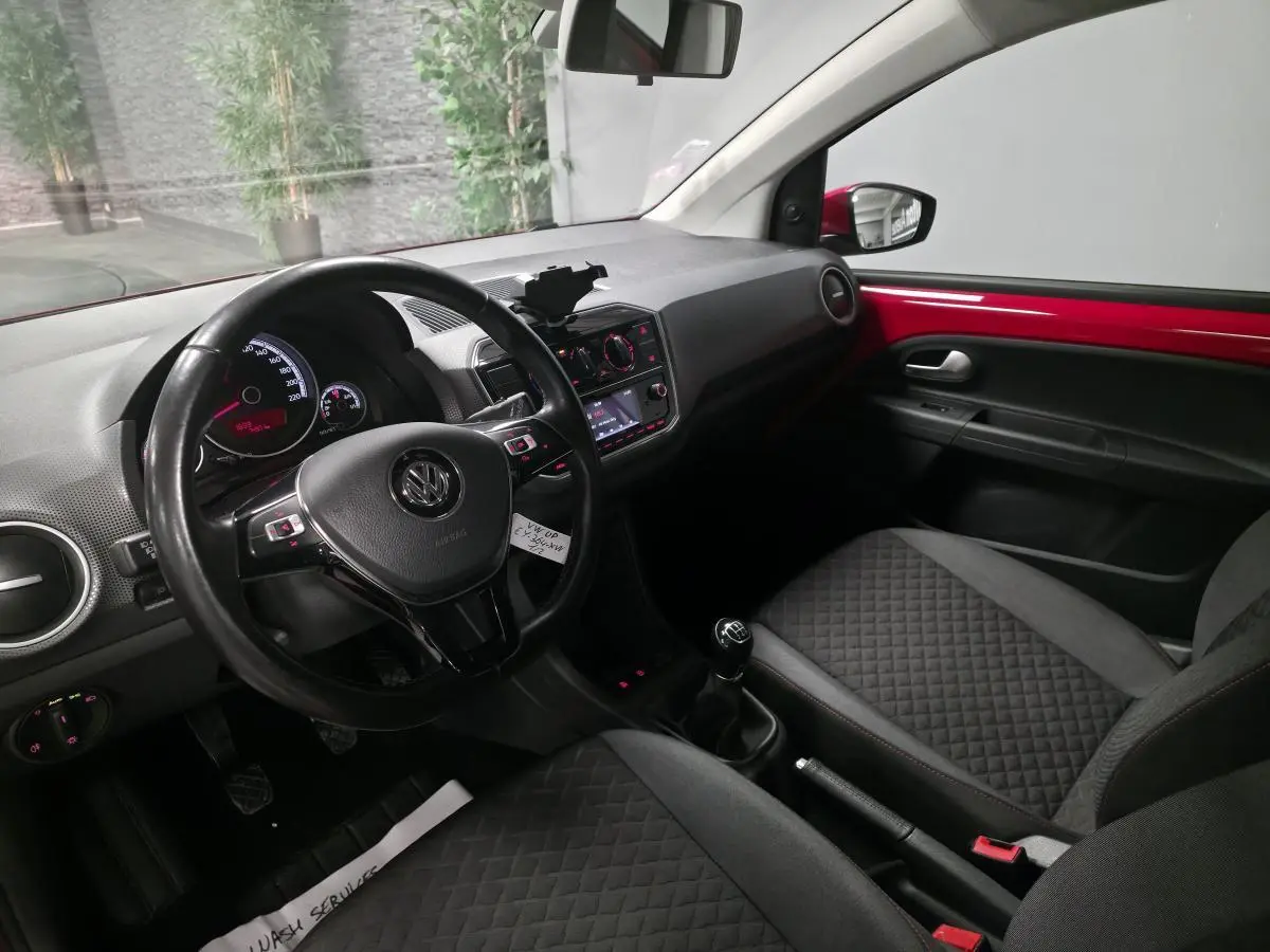Intérieur avant droit d'une Volkswagen Up rouge, volant cuir multifonctions et sellerie tissu avec surpiqûres rouges.
