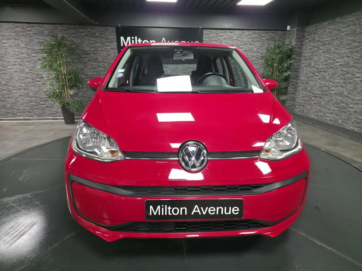 Vue frontale d'une Volkswagen Up rouge vif avec logo VW centré et plaque "Milton Avenue" en intérieur showroom.