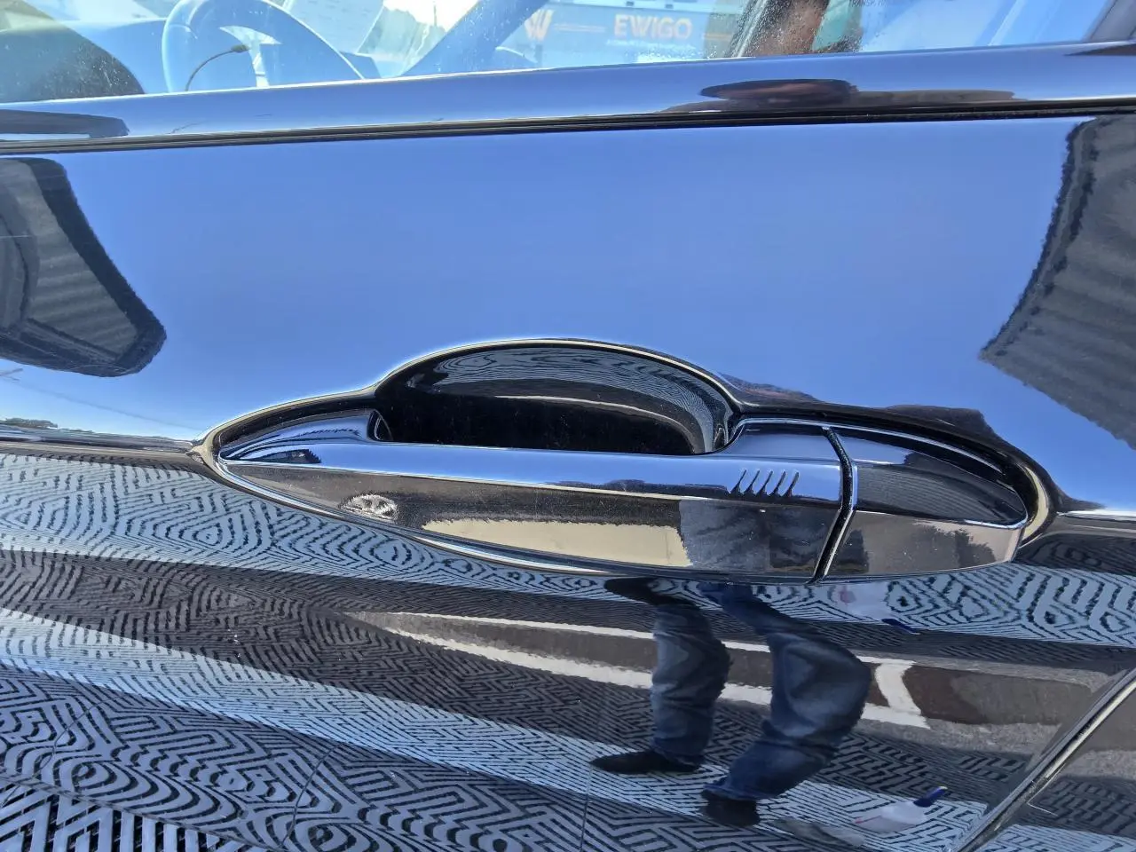 Poignée de porte noire brillante du BMW X1 F48 2020, reflet du sol et d'une personne visible sur la carrosserie noire.