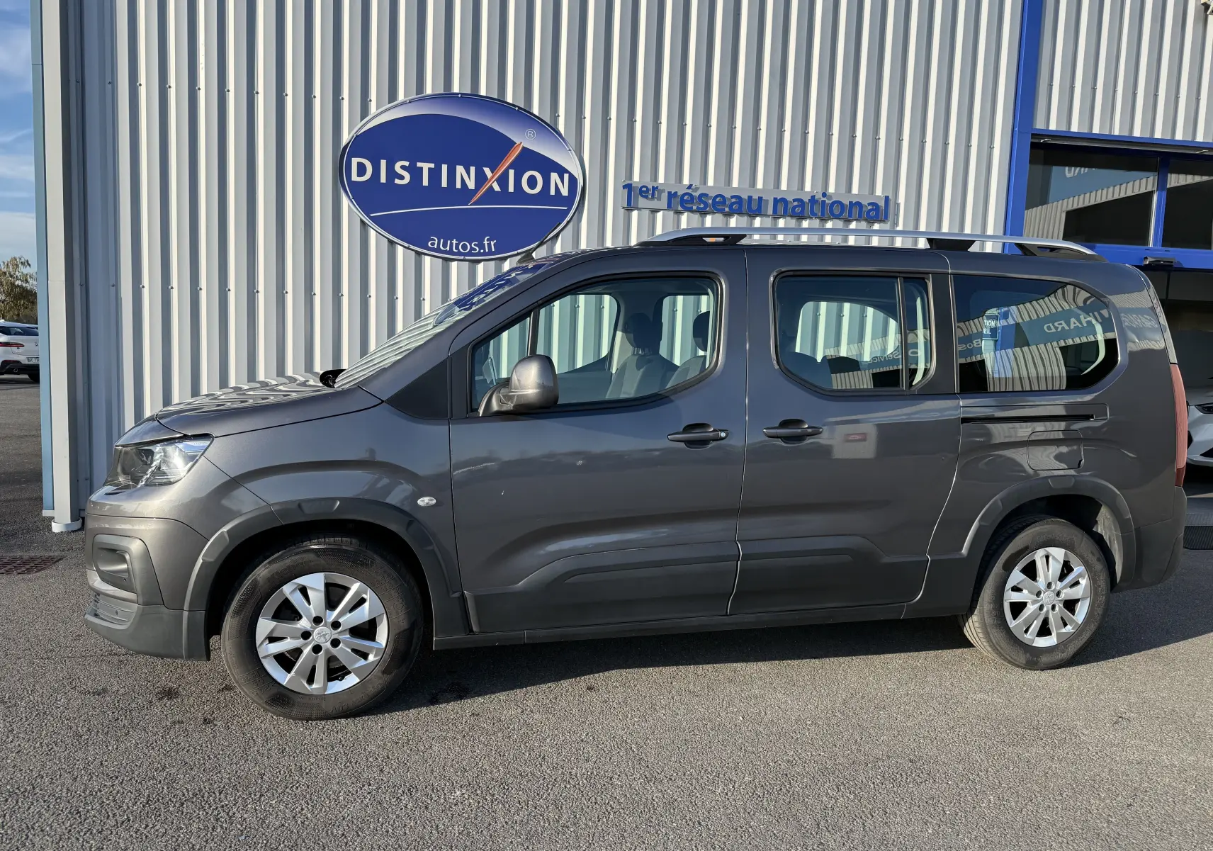 Vue de profil côté gauche d’un Peugeot Rifter gris platinium 7 places garé devant un bâtiment avec enseigne Distinxion.