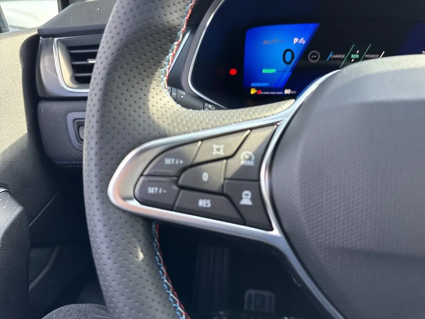 Gros plan sur le volant perforé côté gauche avec commandes multifonctions et tableau de bord numérique bleu du Renault Symbioz blanc nacré.