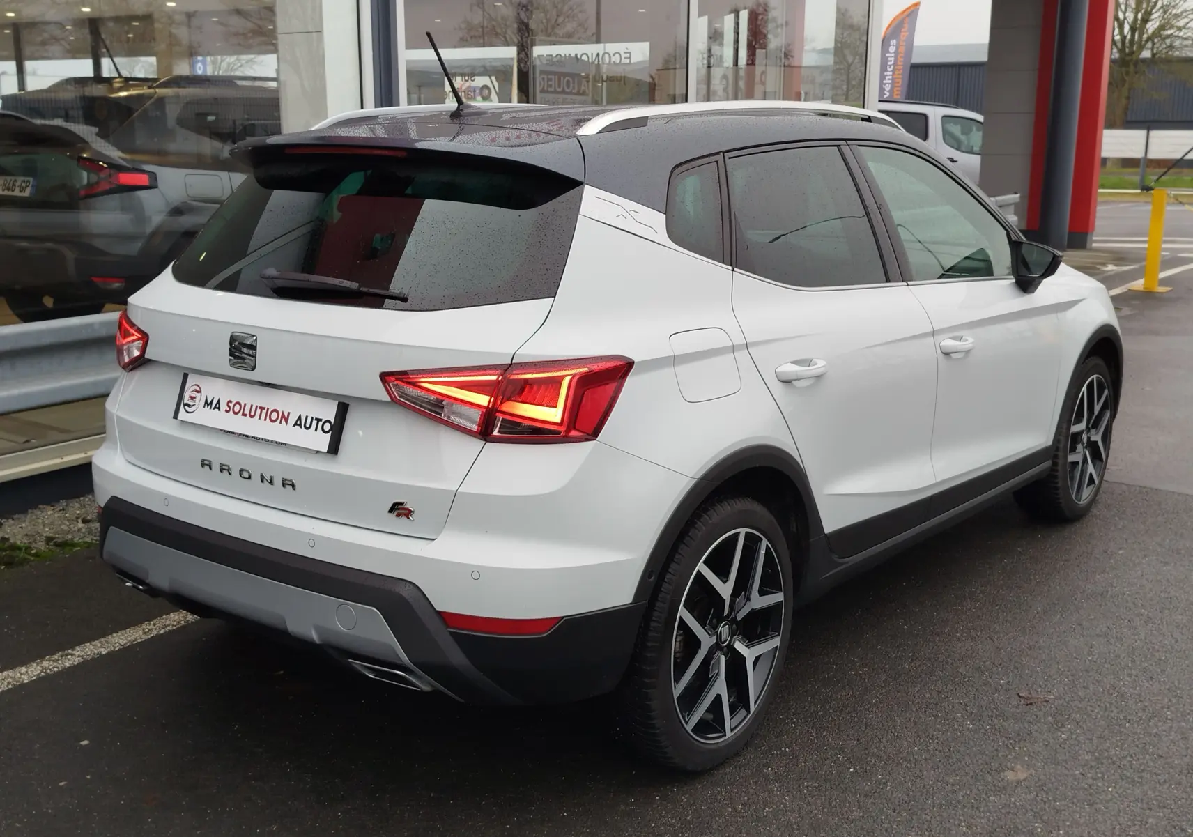Vue 3/4 arrière droite d'un SEAT Arona blanc avec toit gris magnétique, feux arrière allumés et jantes alliage noires.
