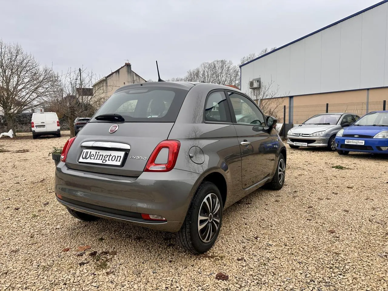 Fiat 500 gris foncé vue 3/4 arrière droit sur un sol gravillonné avec plusieurs voitures en arrière-plan.