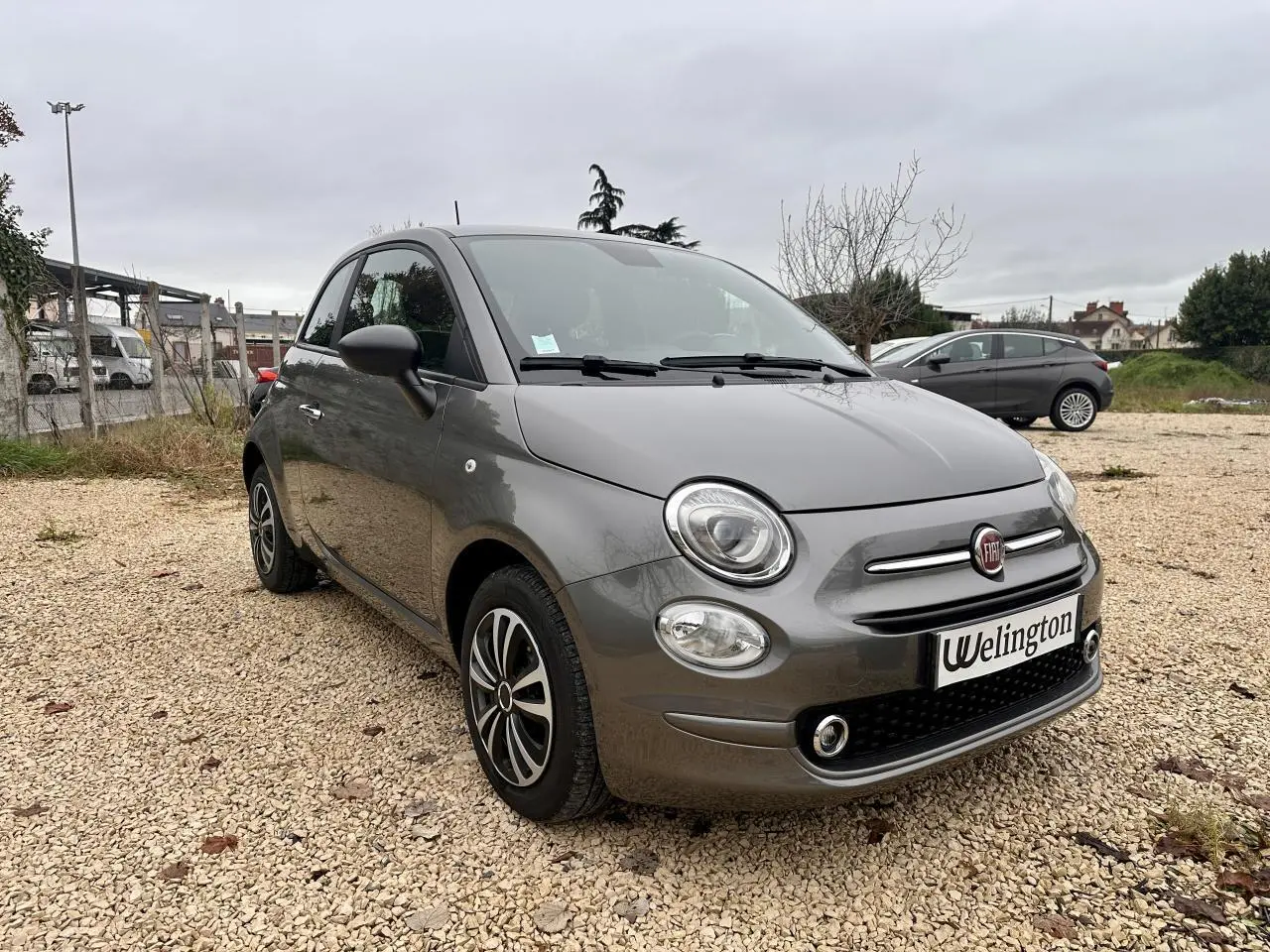 Fiat 500 gris foncé vue 3/4 avant droit sur un sol gravillonné avec jantes alliage et phares ronds.