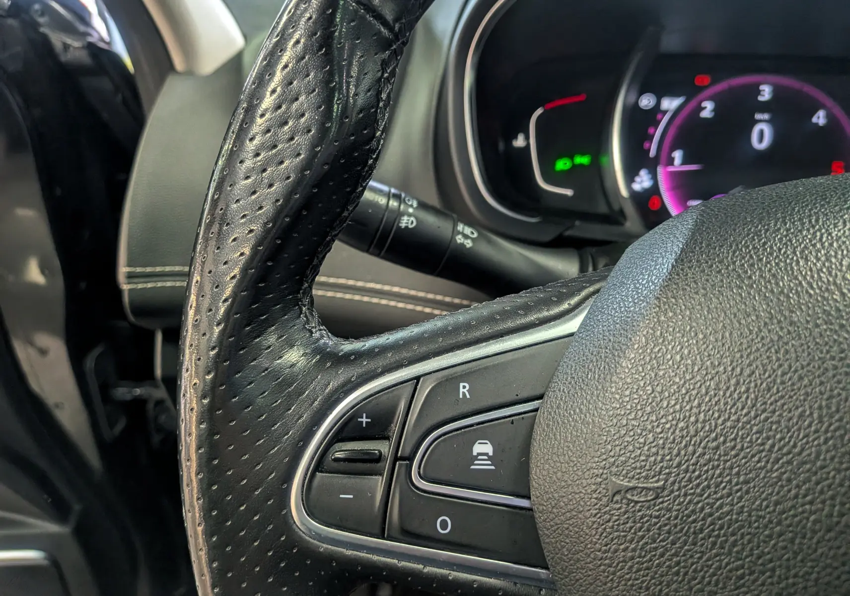 Gros plan sur le volant en cuir noir perforé du Renault Scénic IV, avec commandes intégrées et tableau de bord numérique en arrière-plan.