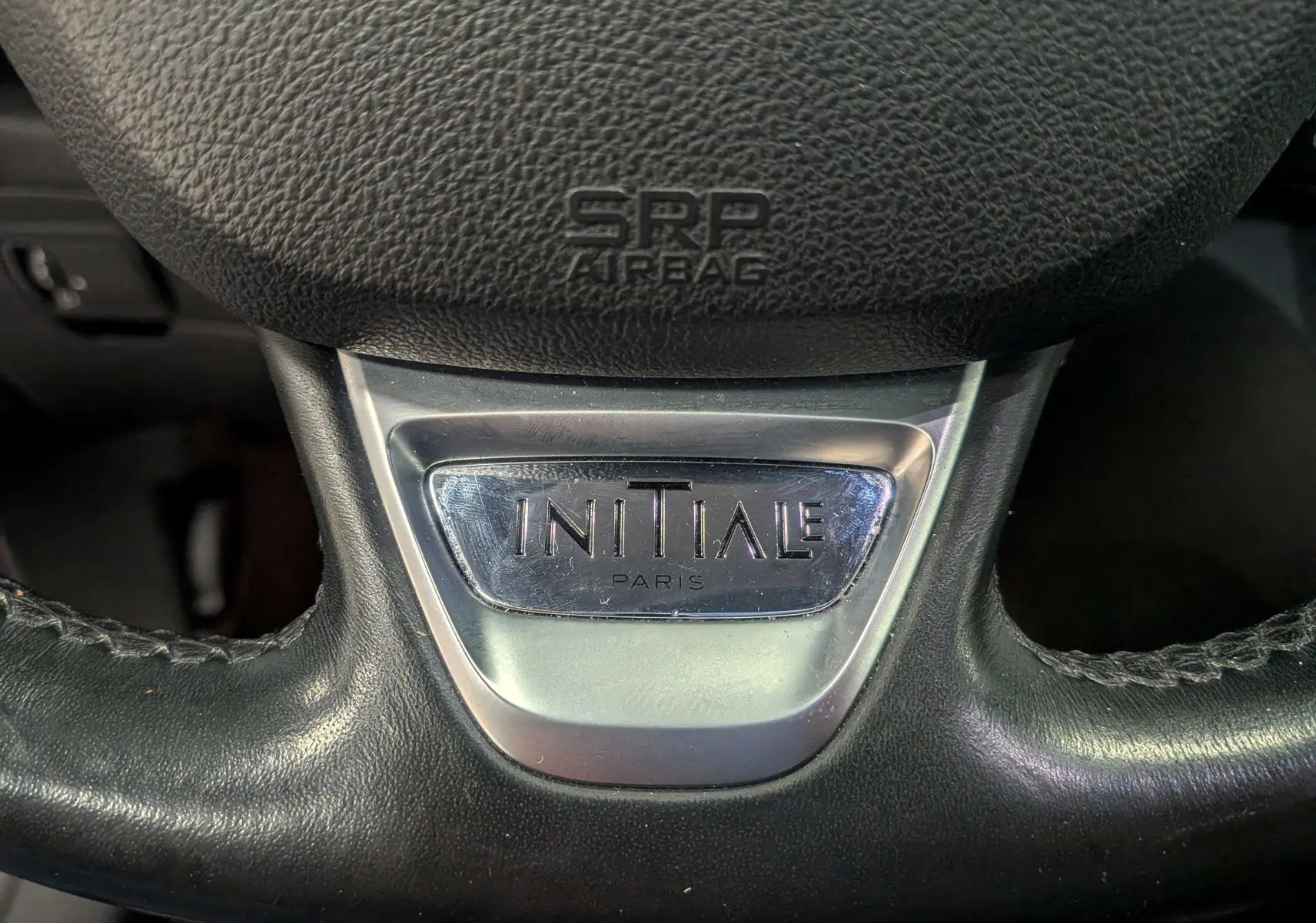 Gros plan sur le volant en cuir noir du Renault Scénic IV Initiale Paris, avec logo métallique "Initiale Paris" au centre.