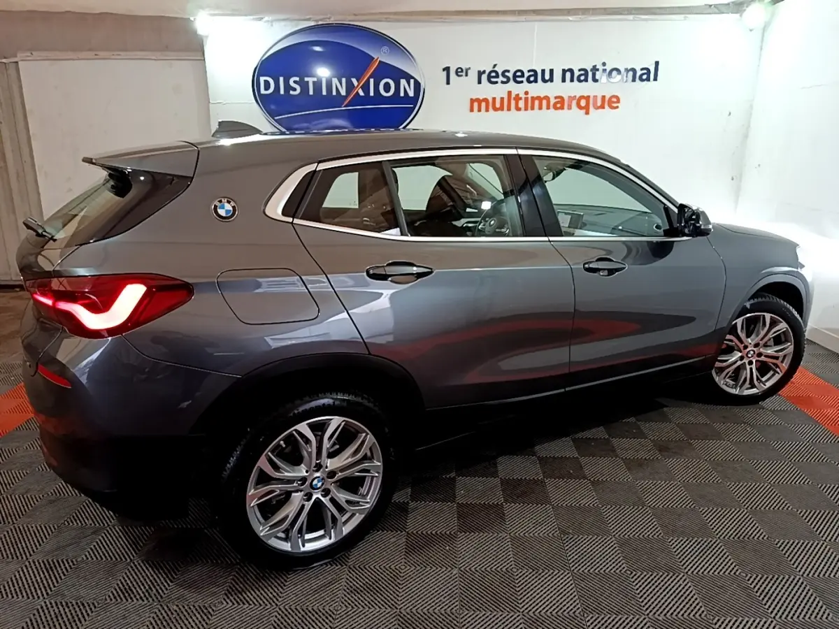 BMW X2 sDrive18i gris foncé vue de profil droit en intérieur avec feux arrière allumés et jantes alliage visibles.