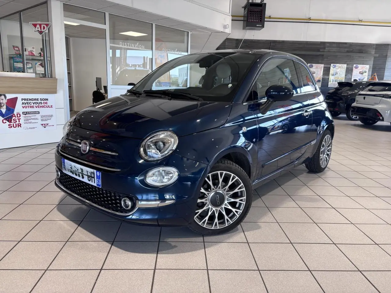 Vue 3/4 avant d'une Fiat 500 bleu foncé avec jantes alliage et toit panoramique en showroom.