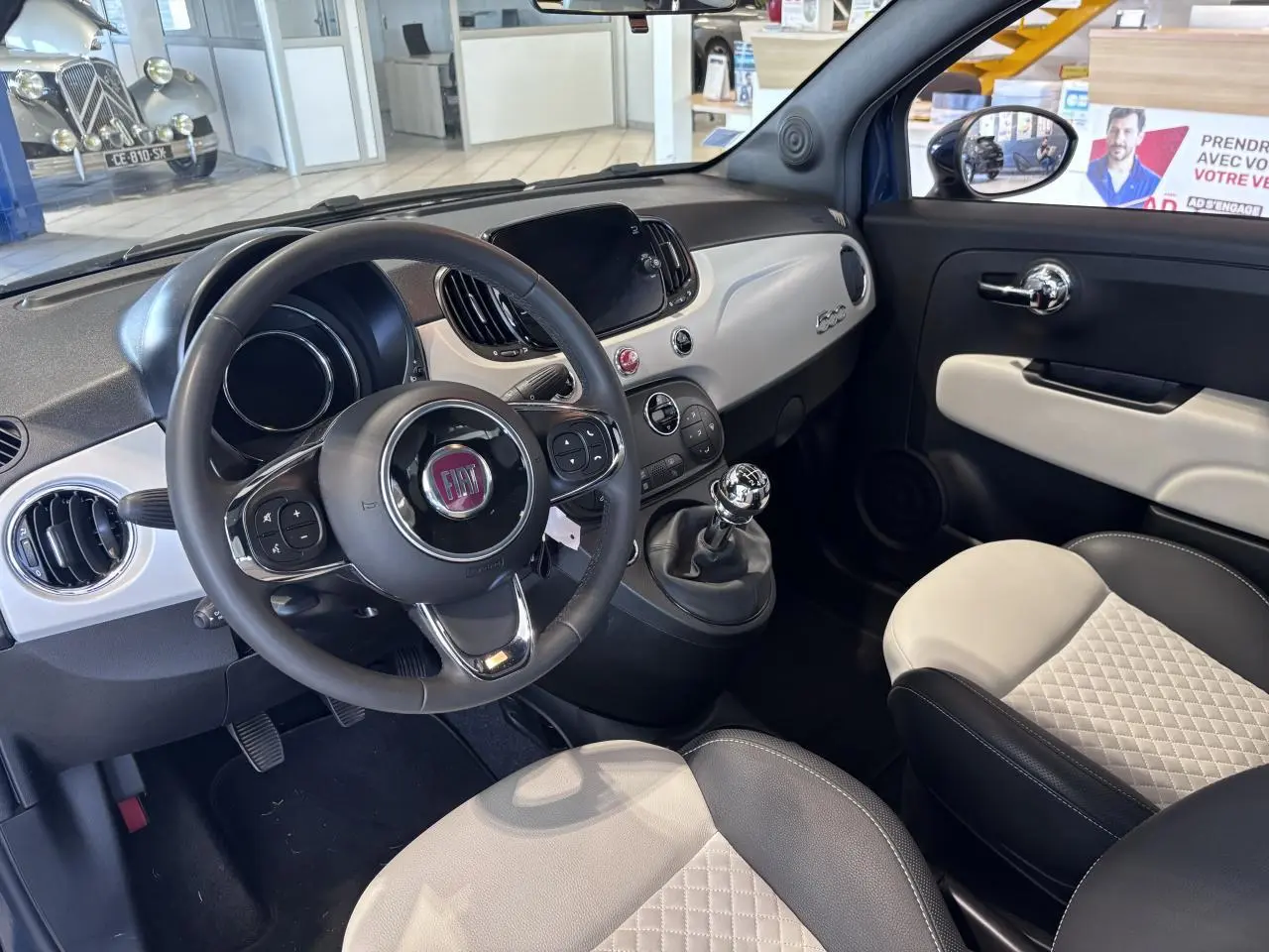 Vue intérieure côté conducteur de la Fiat 500 avec volant simili-cuir, tableau de bord blanc et sièges bicolores matelassés.