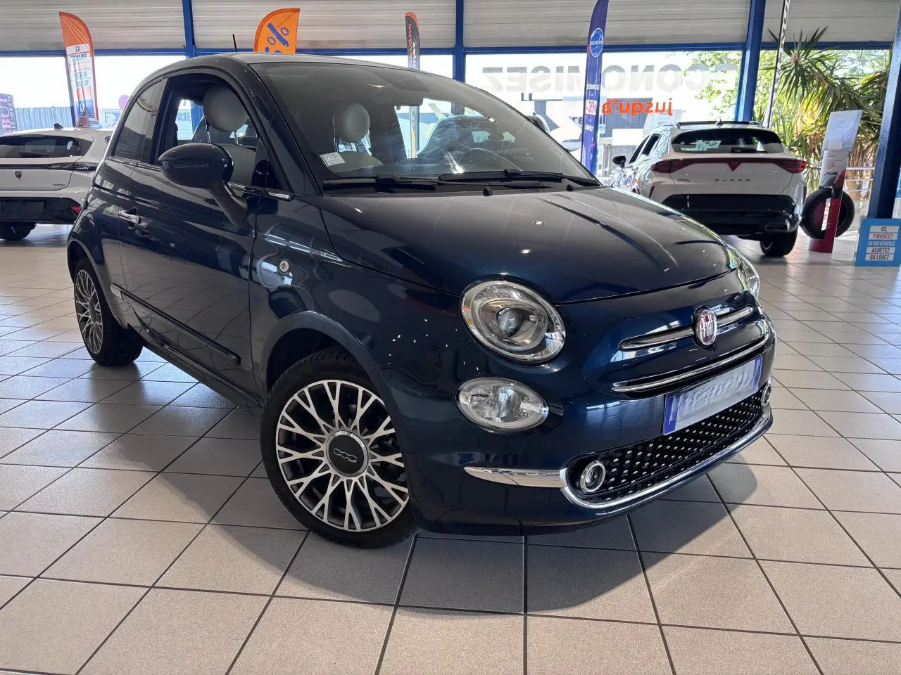 Fiat 500 bleu foncé vue 3/4 avant droit en showroom, avec jantes alliage et calandre chromée.