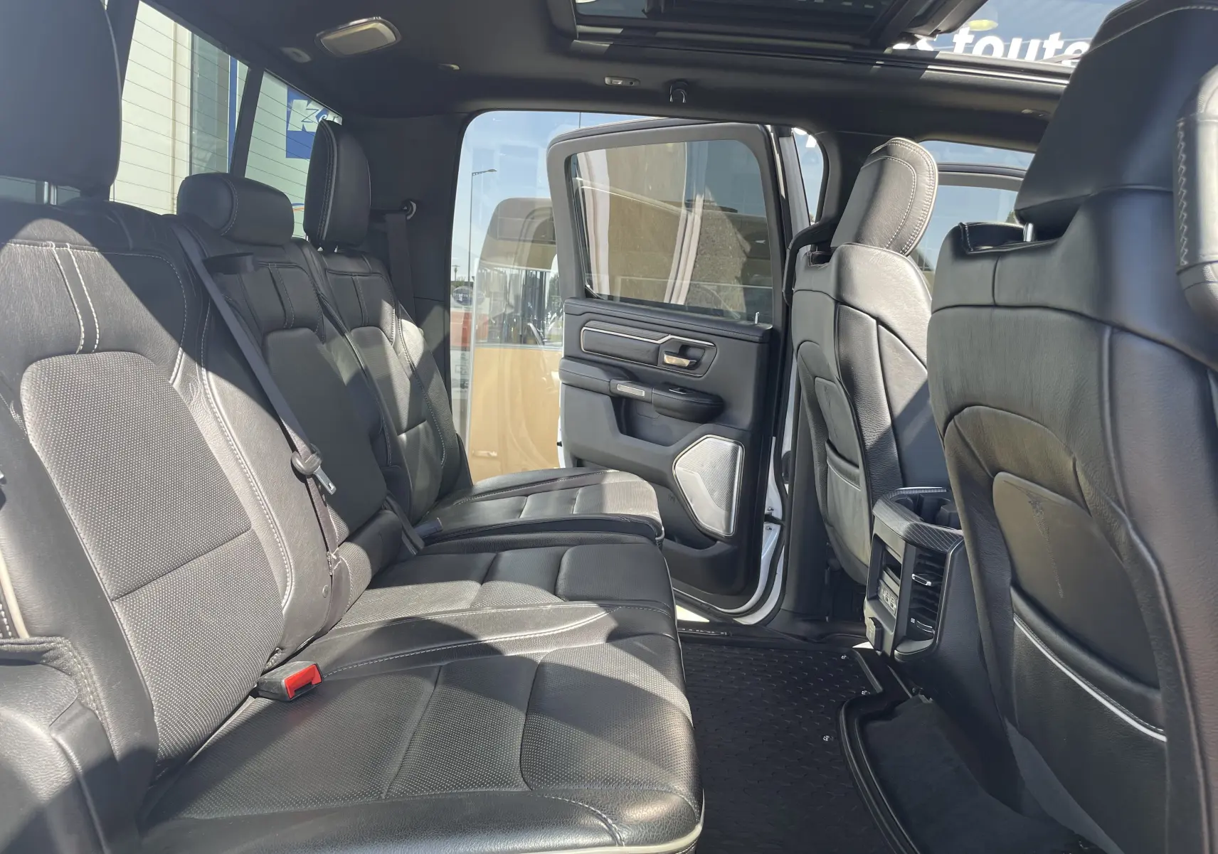 Vue intérieure arrière du Dodge RAM 1500 blanc nacré, sièges en cuir noir et porte arrière ouverte laissant entrer la lumière naturelle.