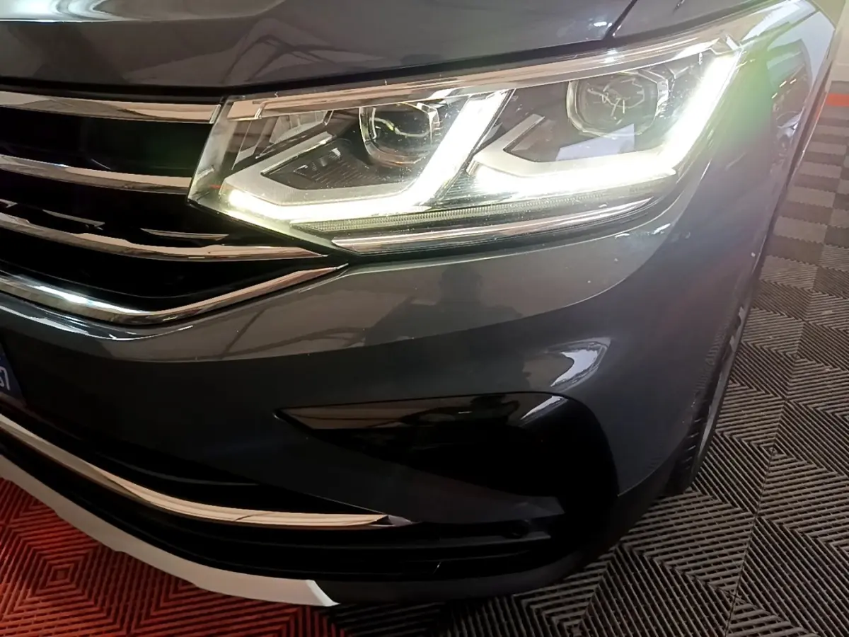 Gros plan sur le phare avant droit allumé d'un Volkswagen Tiguan gris clair avec calandre chromée visible.